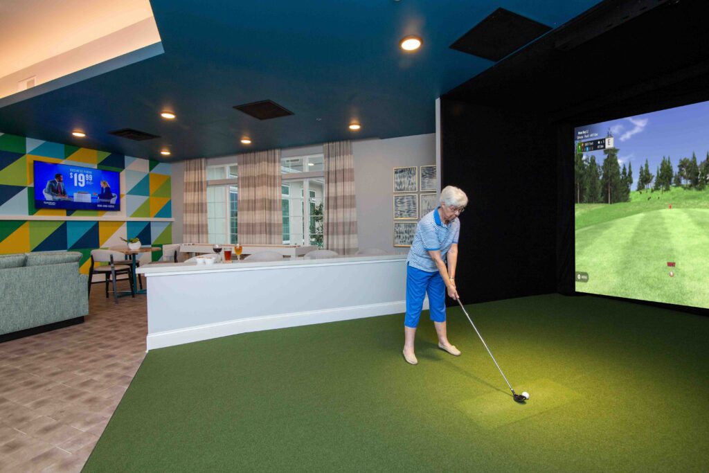 An older white woman wearing a blue shirt and blue pants is playing a golf simulator on the big screen, getting ready to hit the ball at The Atrium of Belleville.