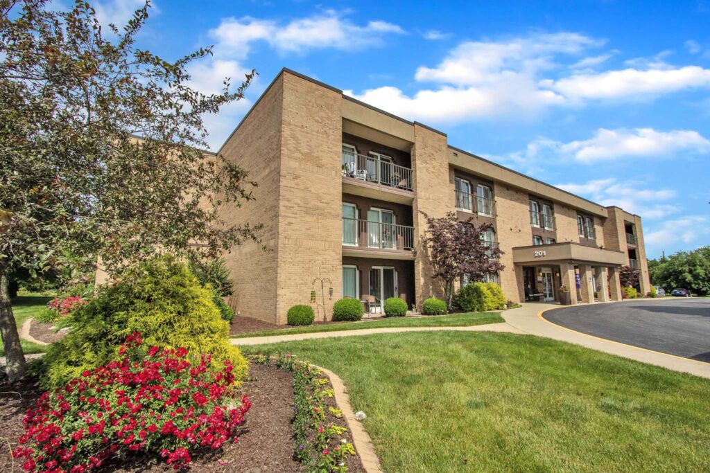 The Atrium of Belleville main building with a driveway leading to it, green grass, and flowering bushes.