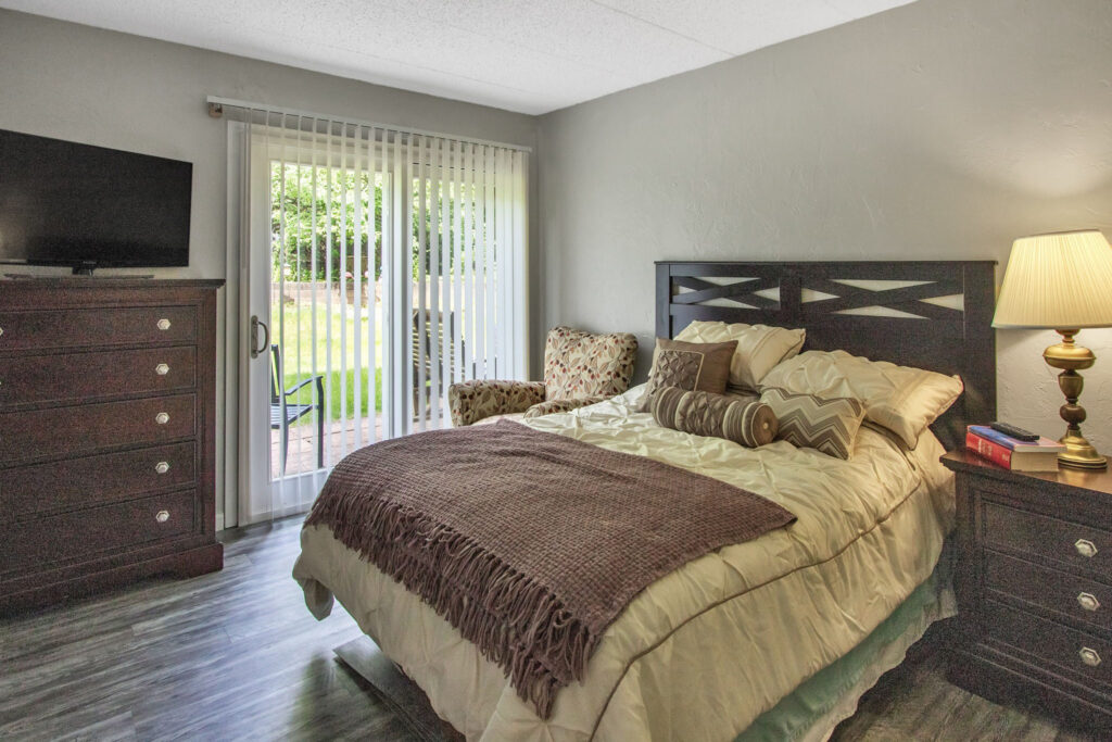 A bedroom with a made-up bed with gold sheets and pillows and a brown throw blanket, a bedside table with a lamp and books on it, a sofa chair, a dresser, and a TV on top of it at the atrium of Belleville.