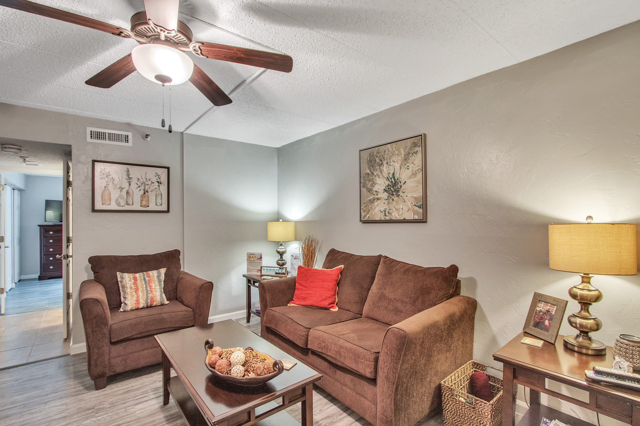 A living room with a brown couch and sofa chair, a coffee table, a ceiling fan, and a side table with a lamp and a framed photograph at The Atrium of Belleville.