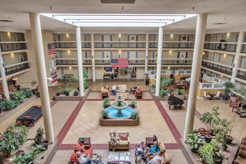 An aerial view of The Atrium of Belleville lobby, with a fountain in the middle, a pool table to the left, a piano to the right, and people sitting on couches and sofa chairs around the lobby.