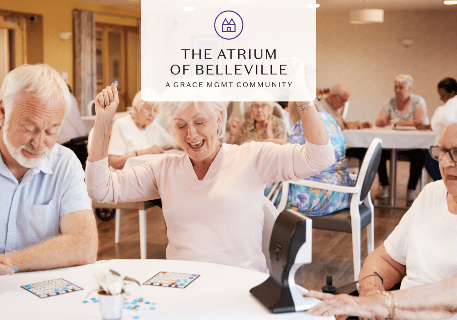 Elderly woman smiles and raises her arms in celebration at a bingo table in a senior living community.