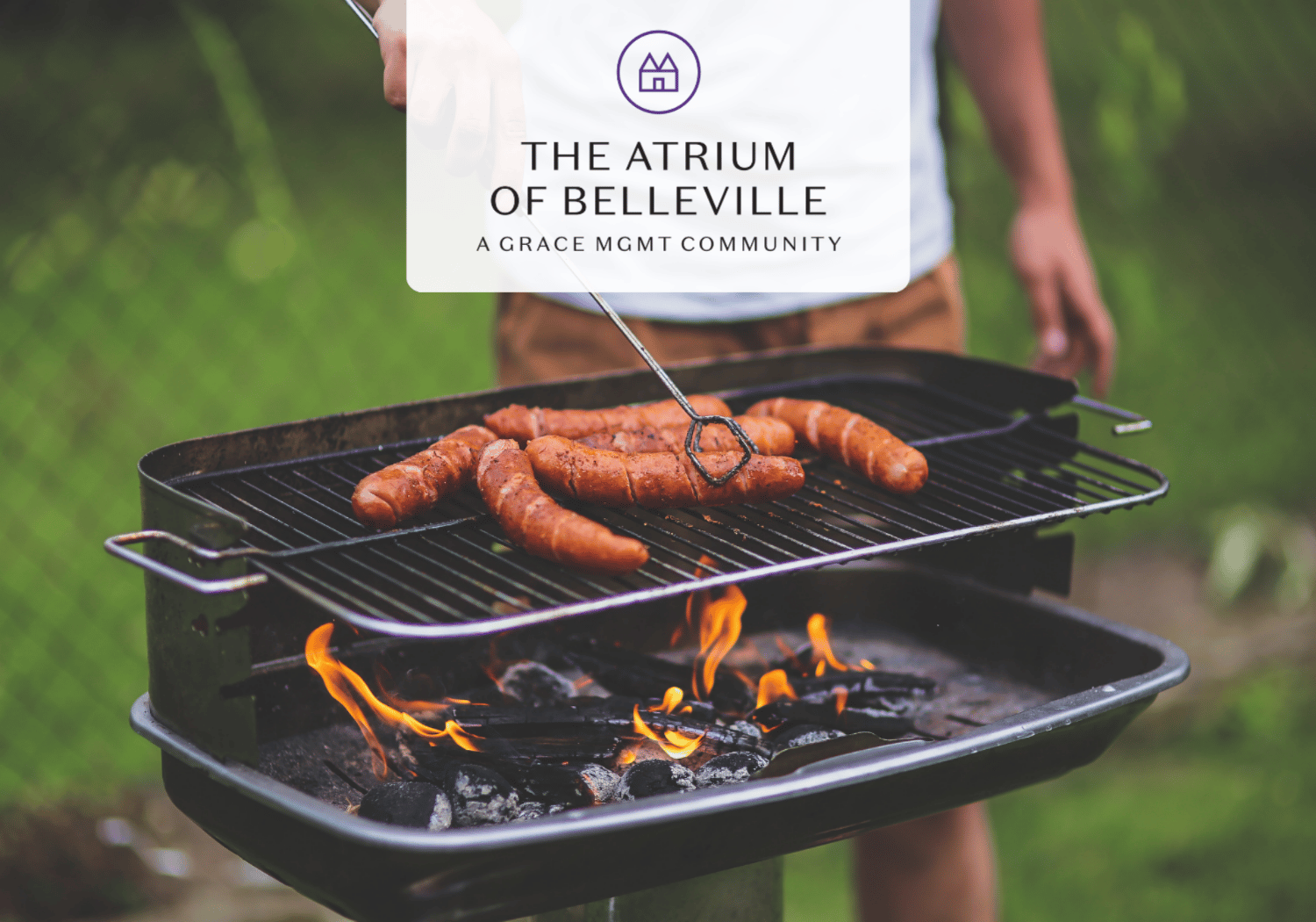 Sausages grilling on a charcoal BBQ outdoors with a blurred person in the background and a logo overlay reading 'The Atrium of Belleville'