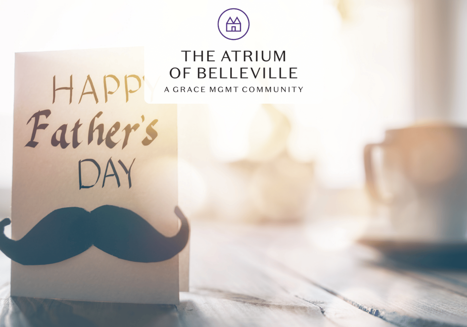 Father's Day card with a blue mustache cutout on a wooden table, soft backlight, and a blurred mug in the background; logo reads The Atrium of Belleville at the top (overlay).