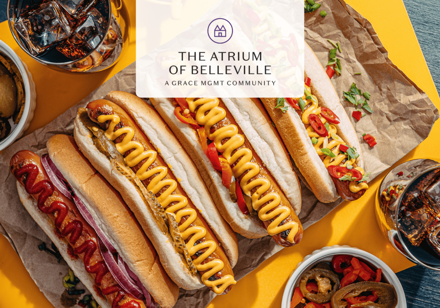 Assorted hot dogs with mustard, onions, and peppers on parchment over a bright yellow surface, with drinks nearby and a translucent logo overlay reading 'The Atrium of Belleville.'